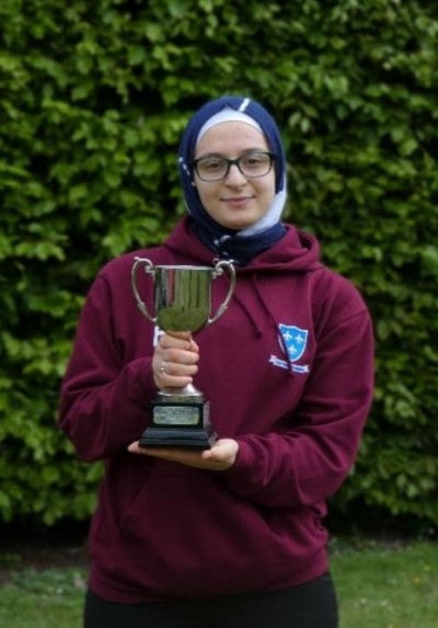 A girl wearing school top and headscarf holds a silver trophy in front of a green hedge.