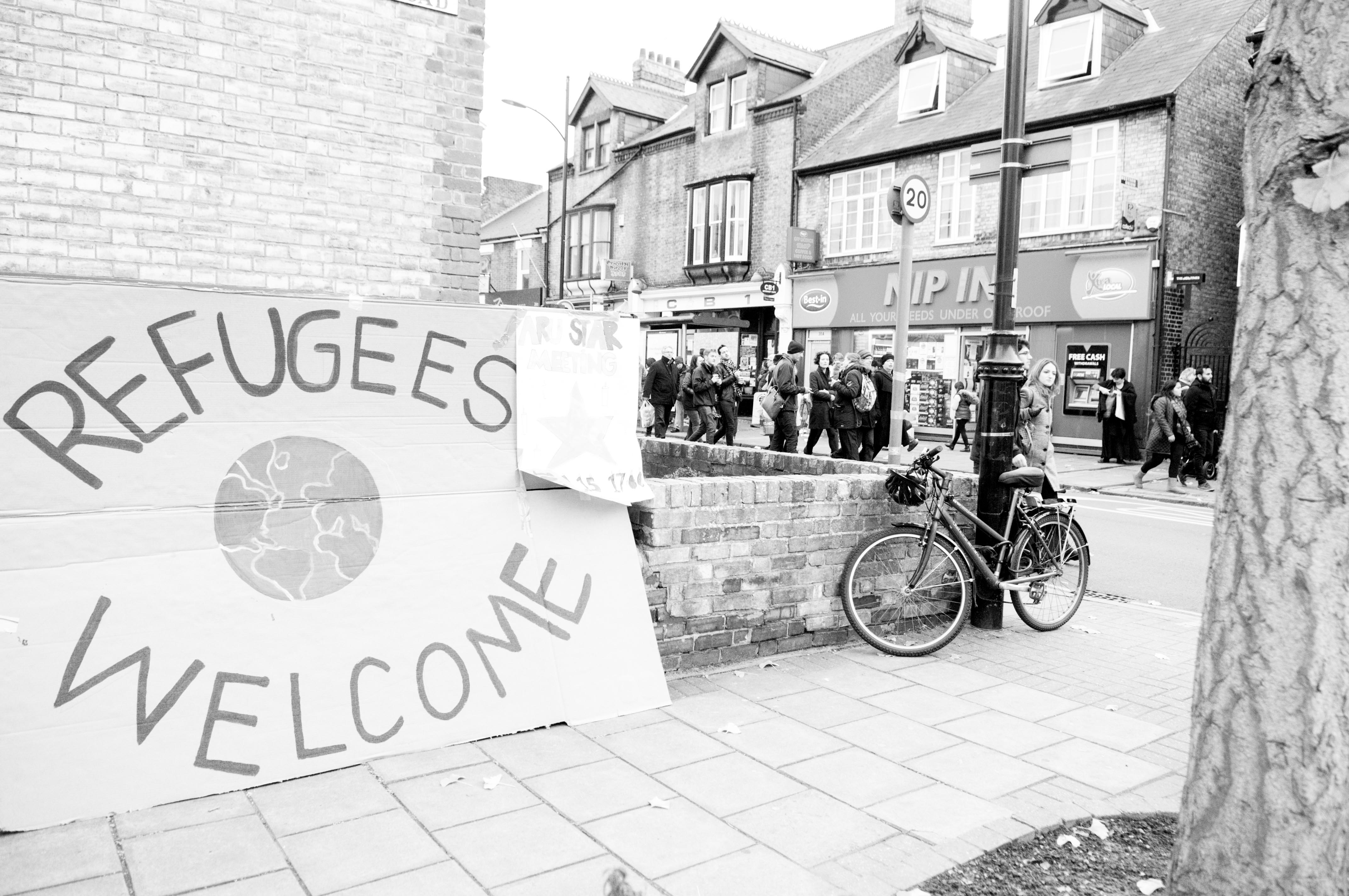 A march along Mill Road supporting refugees and asylum seekers in Cambridge, with a large sign leaning against a wall that says 'Refugees welcome'. Photo copyright Chris Cellier Photography www.chriscellier.com and used with permission.
