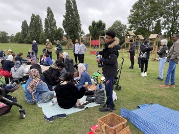 Volunteers and families picnicking on a green, seated in groups with playground equipment in the background