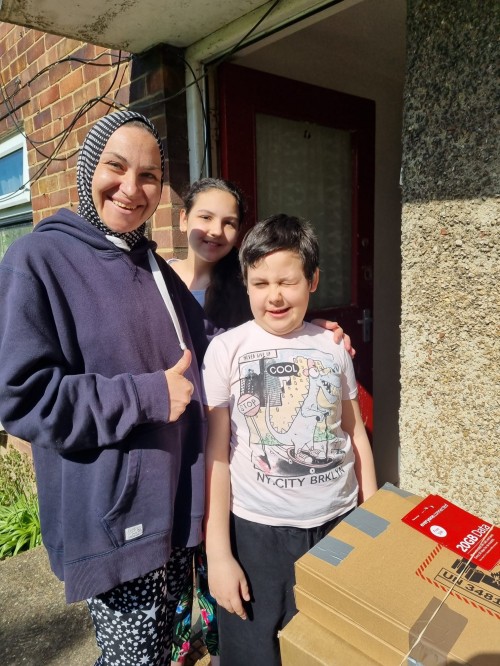 A woman in hoodie and headscarf smiles with her two children at their front door with cardboard boxes marked Fragile
