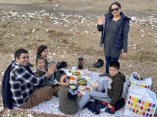 A group of people on a beach, with a man sne four children sat on a blanket waving, while a woman stands behind them also waving