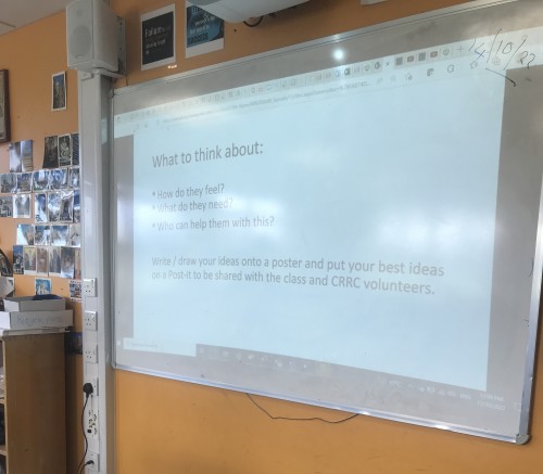 An interactive whiteboard showing a slide with the text "What to think about: how do they feel?; what do they need?; who can help them with this? Write/draw your ideas onto a poster and put your best ideas on a Post-it to be shared with the class and CRRC volunteers" 