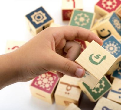 A hand holding a wooden block decorated with Arabic script, with other wooden blocks in the background with traditional islamic tile designs.